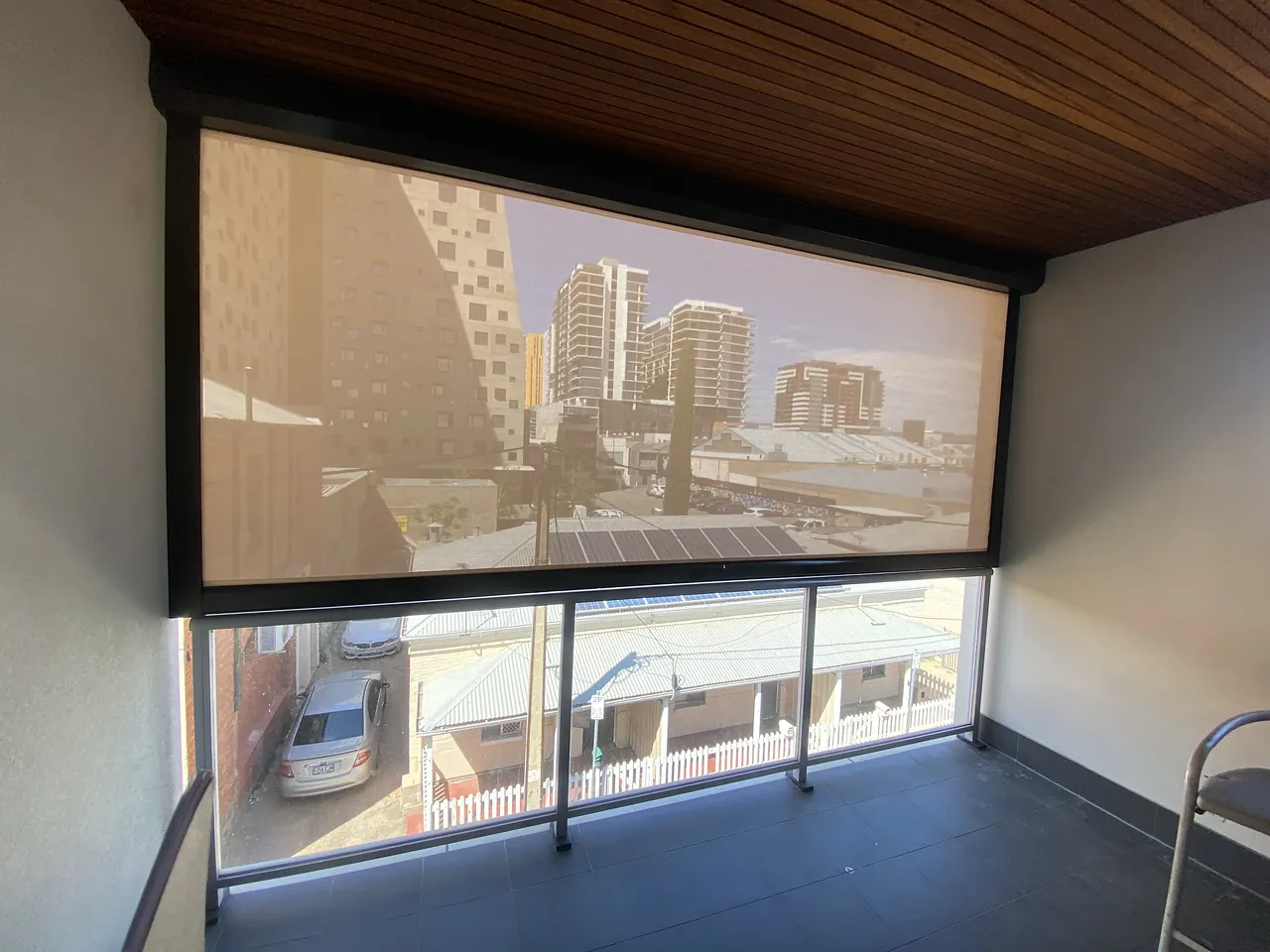 Sheer zip blind lowered on an apartment balcony, city towers visible through the fabric