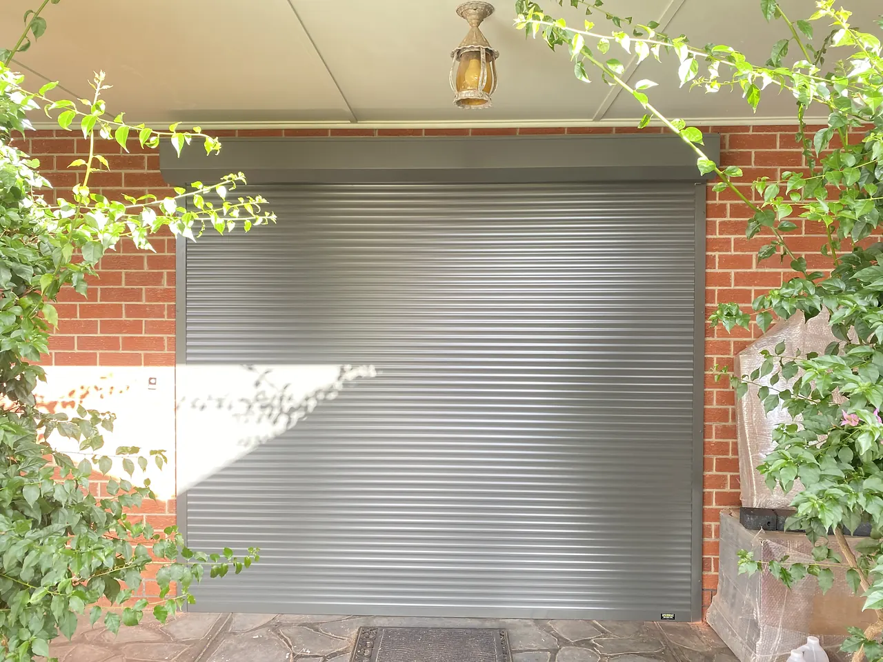 Silver roller shutter fitted inside a brick porch framed by climbing plants