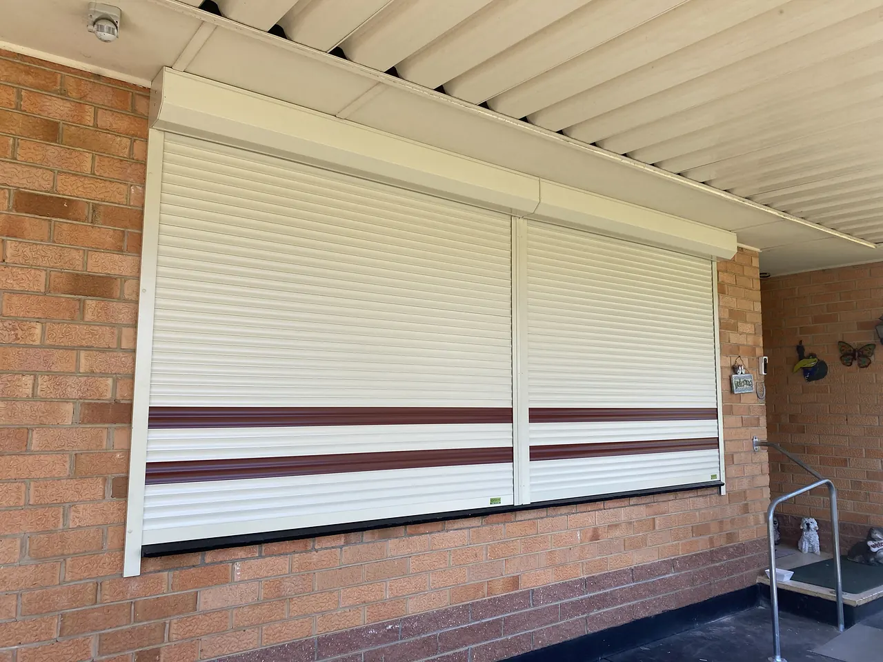 Two cream roller shutters with red accent stripes on a brick home