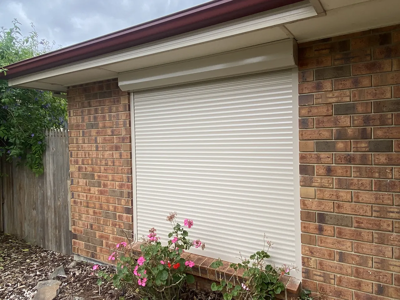 White roller shutter on a red brick home above a flower bed